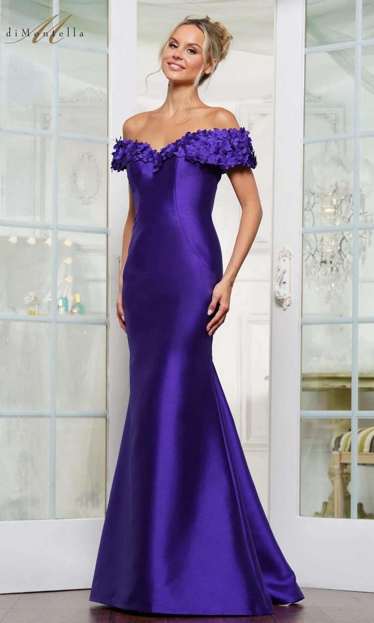 A woman stands indoors in front of glass doors, wearing the Rina Di Montella RD3251 long formal dress featuring a floor-length satin crepe draped skirt and floral appliqué along the off-the-shoulder neckline.